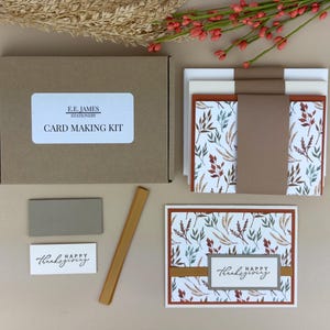May include: A card making kit in a brown cardboard box labeled E.E. JAMES STATIONERY CARD MAKING KIT. Includes stacks of cards with autumn leaf patterns, brown paper bands, a gold strip, a gray rectangle, and a white label reading HAPPY THANKSGIVING.