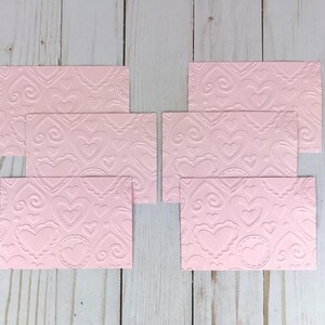 Op de afbeelding: Roze ge&euml;mbosseerd karton met een herhalend hart- en krulpatroon. De kaarten zijn rechthoekig en in een groep gerangschikt. Het ontwerp bevat verschillende maten harten en decoratieve randen.