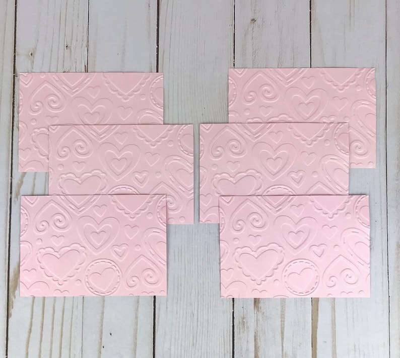 Op de afbeelding: Roze ge&euml;mbosseerd karton met een herhalend hart- en krulpatroon. De kaarten zijn rechthoekig en in een groep gerangschikt. Het ontwerp bevat verschillende maten harten en decoratieve randen.
