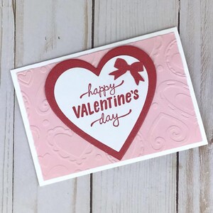 Op de afbeelding: Een Valentijnsdagkaart met een rood hartontwerp en een strik. De kaart heeft een witte rand en een roze achtergrond met reli&euml;f hartpatronen. De woorden "happy Valentine's day" zijn in rood schrift geschreven.