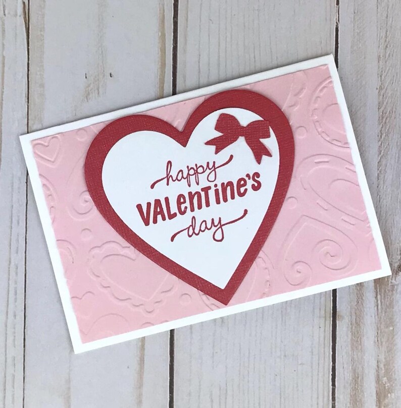 Op de afbeelding: Een Valentijnsdagkaart met een rood hartontwerp en een strik. De kaart heeft een witte rand en een roze achtergrond met reli&euml;f hartpatronen. De woorden "happy Valentine's day" zijn in rood schrift geschreven.