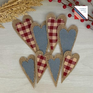 May include: A collection of heart-shaped burlap ornaments with red and white, and blue and white checkered fabric centers. The ornaments are arranged on a light-colored surface, with a sprig of berries and wheat stalks in the background. The text "Celebrate America 2026" is visible.