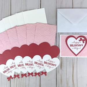 Op de afbeelding: Een Valentijnsdagkaartenset met rode en witte hartvormige kaarten met de tekst "happy VALENTINE's day". De set bevat ook roze reli&euml;fkaarten, witte enveloppen en kleine rode strikjes.