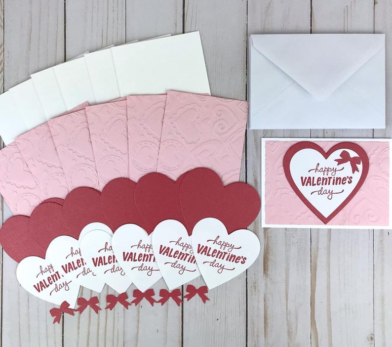 Op de afbeelding: Een Valentijnsdagkaartenset met rode en witte hartvormige kaarten met de tekst "happy VALENTINE's day". De set bevat ook roze reli&euml;fkaarten, witte enveloppen en kleine rode strikjes.