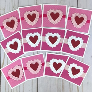 May include: A collection of eleven square greeting cards featuring a dark red heart centered on a white or light pink ornate die-cut heart frame. Each card has a horizontal ribbon strip across the center and a pink background with a subtle dot pattern.