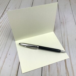Pu&ograve; includere: Un biglietto d'auguri piegato color bianco sporco posizionato verticalmente, con una penna a sfera nera e argento appoggiata orizzontalmente sulla met&agrave; inferiore, dotata di una clip argentata e di una punta e un cappuccio lucidi.