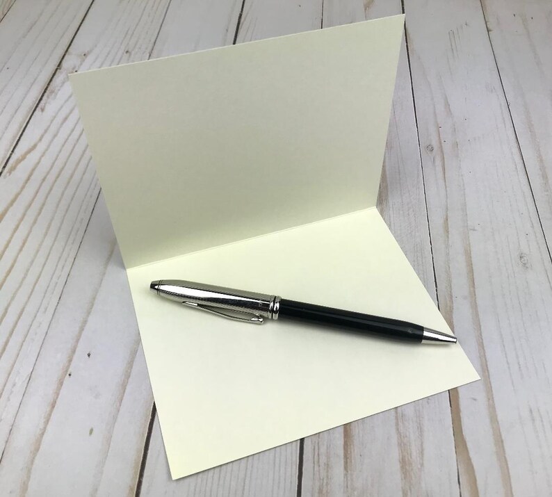 Pu&ograve; includere: Un biglietto d'auguri piegato color bianco sporco posizionato verticalmente, con una penna a sfera nera e argento appoggiata orizzontalmente sulla met&agrave; inferiore, dotata di una clip argentata e di una punta e un cappuccio lucidi.