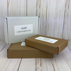 May include: Three card making kits are stacked on a light wood surface. Two are brown, and one is white. Each box has a white label with black text that reads "CARD MAKING KIT". The white box has the text "F.E. JAMES STATIONERS" above the label.