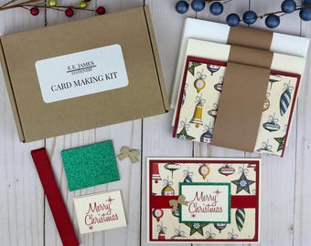 May include: A card making kit in a brown cardboard box labeled E.E. James Stationery. Includes stacks of cream and white envelopes, cards with a vintage Christmas ornament pattern, a red ribbon, a green glitter square, and a Merry Christmas card.
