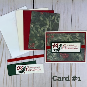 Puede incluir: Un juego de tarjetas navide&ntilde;as que incluye una tarjeta rectangular con un patr&oacute;n de mu&eacute;rdago verde oscuro y bayas rojas, un borde rojo y una etiqueta central blanca con el texto Merry Christmas. Incluye sobres y papel a juego.