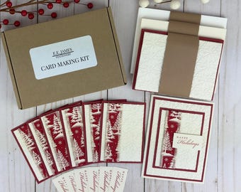 以下が含まれることがあります： E.E. James Stationeryとラベルが貼られた茶色の段ボール箱に入ったカードメイキングキット。エンボス加工が施されたクリーム色のカード、赤と白のクリスマスツリー柄の紙、Happy Holidaysと印刷された小さな長方形のタグが含まれています。