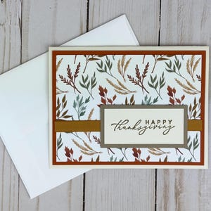 May include: A rectangular greeting card with a white background featuring a pattern of autumn-colored leaves in shades of rust, sage green, and gold. A horizontal gold ribbon crosses the center, topped with a label reading HAPPY thanksgiving.