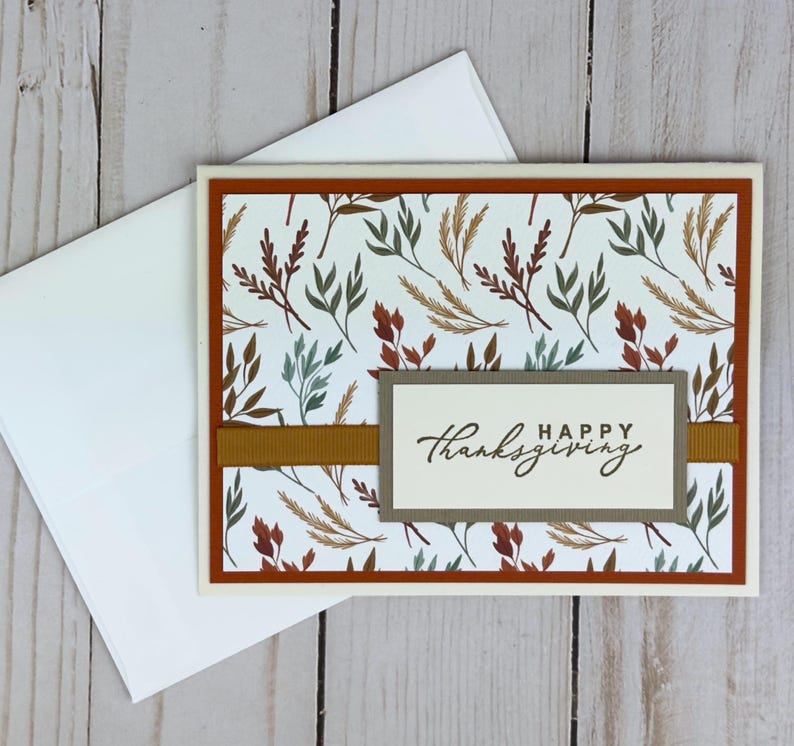 May include: A rectangular greeting card with a white background featuring a pattern of autumn-colored leaves in shades of rust, sage green, and gold. A horizontal gold ribbon crosses the center, topped with a label reading HAPPY thanksgiving.