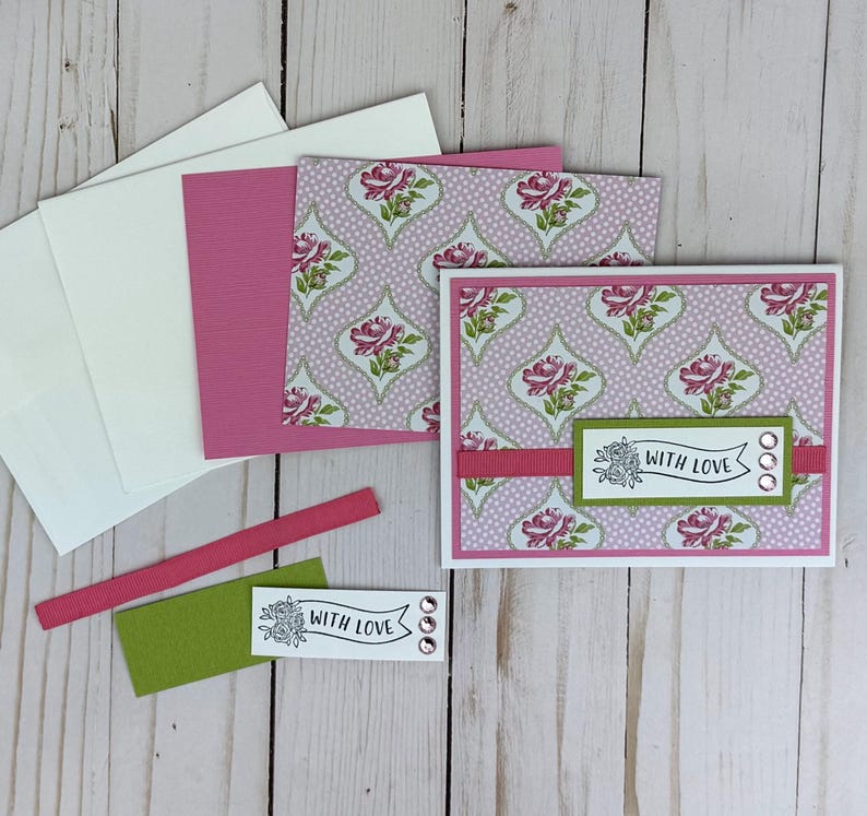 K&ouml;nnte beinhalten: Eine handgefertigte Gru&szlig;karte mit einem rosa-wei&szlig;en Blumenmuster und einem gr&uuml;nen rechteckigen Akzentstreifen. Die Karte zeigt ein wei&szlig;es Banner mit dem Text "WITH LOVE" und drei kleinen rosa Strasssteinen. Passende Umschl&auml;ge enthalten.
