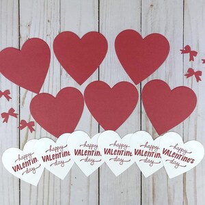 Op de afbeelding: Opstelling van rode papieren harten en kleine rode strikjes op een licht houtnerf oppervlak. Daaronder witte harten met de tekst "happy Valentine's Day" in rode letters.