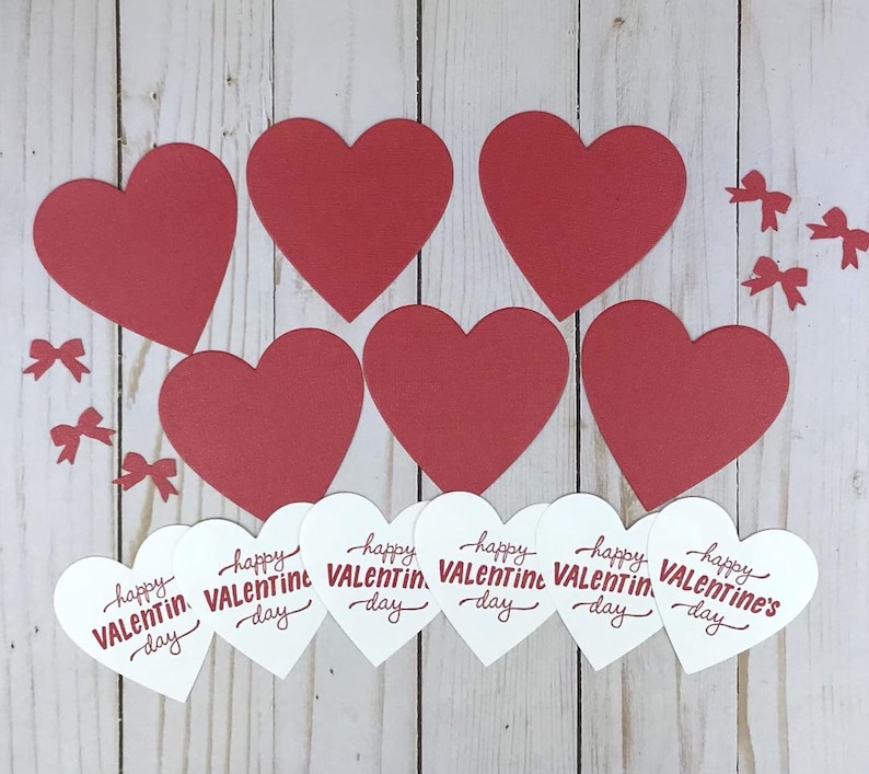 Op de afbeelding: Opstelling van rode papieren harten en kleine rode strikjes op een licht houtnerf oppervlak. Daaronder witte harten met de tekst "happy Valentine's Day" in rode letters.