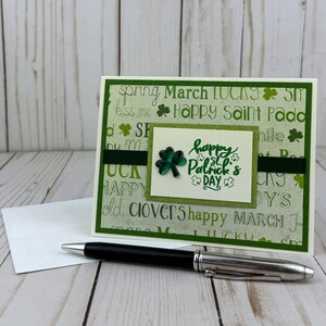 May include: A St. Patrick's Day greeting card with a green and white background featuring words and shamrocks. The card has a green ribbon and a small square with a shamrock and the words "Happy St. Patrick's Day". A black pen and white envelope are in the foreground.
