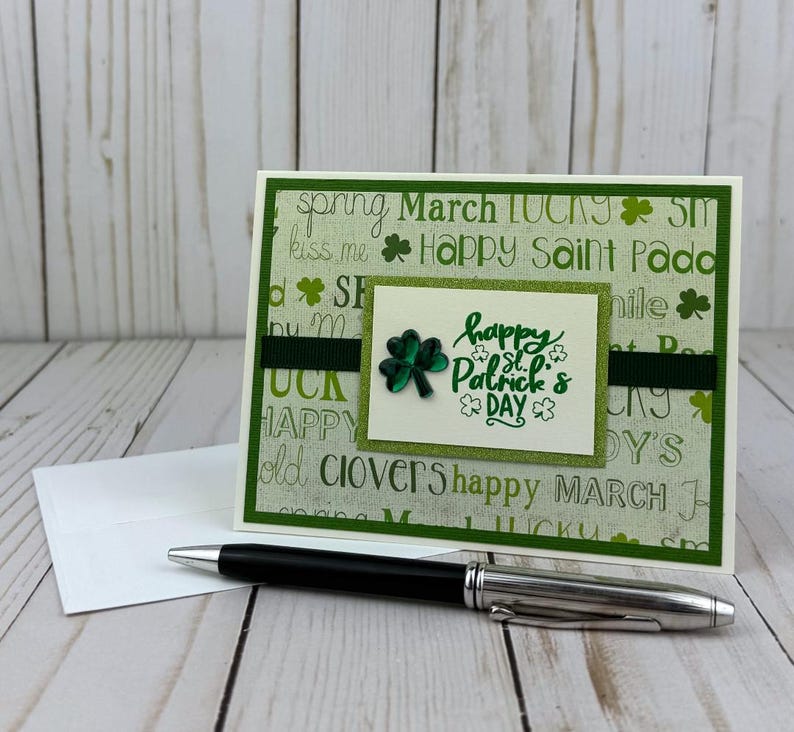 May include: A St. Patrick's Day greeting card with a green and white background featuring words and shamrocks. The card has a green ribbon and a small square with a shamrock and the words "Happy St. Patrick's Day". A black pen and white envelope are in the foreground.