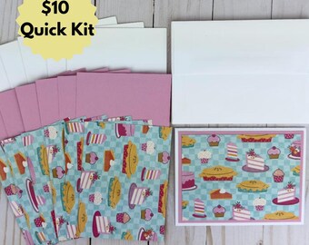 May include: A set of greeting cards and envelopes featuring a light blue and white checkered pattern with various colorful illustrations of cakes, pies, and cupcakes. The cards are paired with solid pink cardstock inserts and white envelopes.