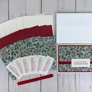 May include: A set of Christmas greeting cards featuring a light blue background with a pattern of green leaves and red berries. Each card has a dark red border and a white rectangular label with the text Merry Christmas in dark red script.
