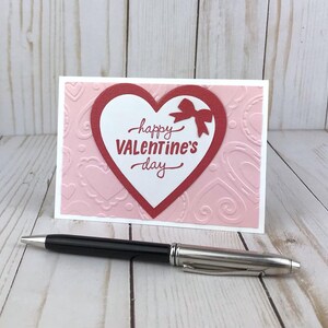Op de afbeelding: Een Valentijnsdagkaart met een rood hartontwerp en de woorden "happy Valentine's day" in rood schrift. De kaart heeft een roze achtergrond met reli&euml;f hartpatronen en een witte rand. Een zwarte en zilveren pen ligt voor de kaart.