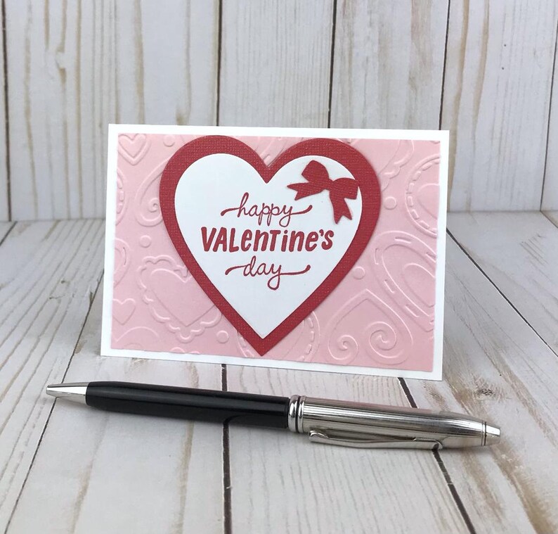 Op de afbeelding: Een Valentijnsdagkaart met een rood hartontwerp en de woorden "happy Valentine's day" in rood schrift. De kaart heeft een roze achtergrond met reli&euml;f hartpatronen en een witte rand. Een zwarte en zilveren pen ligt voor de kaart.