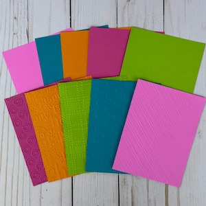 May include: A collection of colorful cardstock paper in various shades of pink, teal, orange, and green. Some cards have embossed patterns, including circles, plaid, and diagonal lines. The cards are arranged on a light wood surface.
