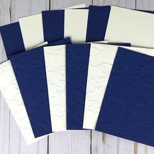 May include: A collection of navy blue and cream-colored cardstock. Some cards are plain, while others feature an embossed floral design. The cards are arranged in a fan shape, showcasing the contrasting colors and textures. Ideal for crafting or stationery.