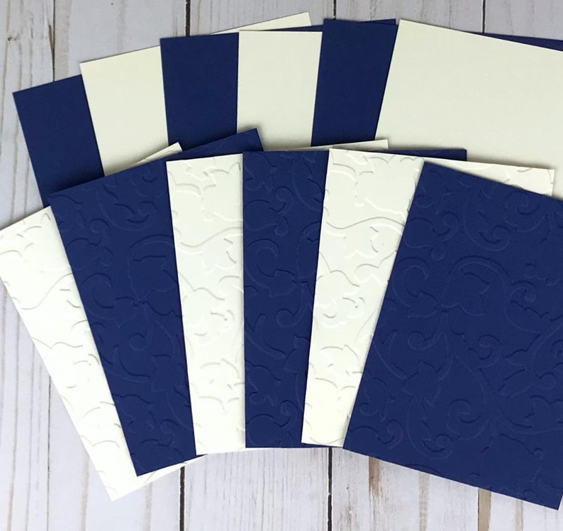 May include: A collection of navy blue and cream-colored cardstock. Some cards are plain, while others feature an embossed floral design. The cards are arranged in a fan shape, showcasing the contrasting colors and textures. Ideal for crafting or stationery.