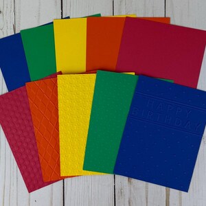May include: A collection of colorful blank greeting cards in shades of red, orange, yellow, green, and blue. Some cards have textured patterns, while one blue card features the embossed text "HAPPY BIRTHDAY".