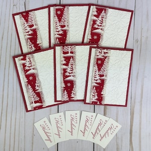 Puede incluir: Cinco tarjetas de felicitaci&oacute;n rectangulares con cartulina color crema en relieve, bordes rojos y una franja vertical de tela roja con pinos y copos de nieve blancos. Debajo de las tarjetas hay seis etiquetas rectangulares con Happy Holidays.