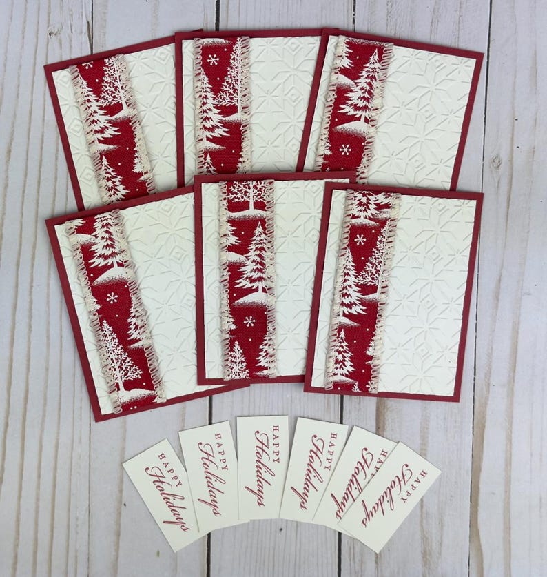 Puede incluir: Cinco tarjetas de felicitaci&oacute;n rectangulares con cartulina color crema en relieve, bordes rojos y una franja vertical de tela roja con pinos y copos de nieve blancos. Debajo de las tarjetas hay seis etiquetas rectangulares con Happy Holidays.