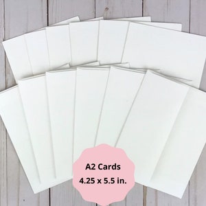 May include: A collection of plain white A2 envelopes and cards arranged in a fan shape. A pink scalloped label in the foreground displays the black text A2 Cards 4.25 x 5.5 in. The stationery items are rectangular with clean edges and a smooth finish.