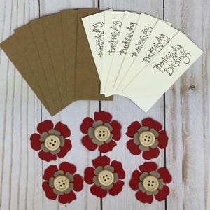 Pu&ograve; includere: Un set di sei etichette rettangolari color crema con il testo "thanksgiving Blessings" stampato in nero, sei etichette in carta kraft marrone e sei fiori di carta a strati rossi e beige, ciascuno con un bottone in legno al centro.