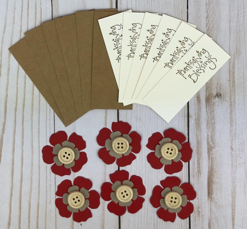 Pu&ograve; includere: Un set di sei etichette rettangolari color crema con il testo "thanksgiving Blessings" stampato in nero, sei etichette in carta kraft marrone e sei fiori di carta a strati rossi e beige, ciascuno con un bottone in legno al centro.