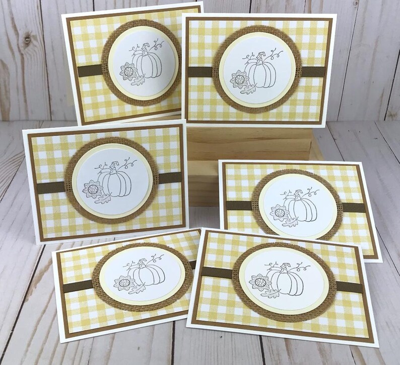 May include: A collection of six rectangular greeting cards featuring a yellow and white gingham pattern. Each card has a central circular white medallion with a black line drawing of a pumpkin and a sunflower, framed by a textured burlap border.