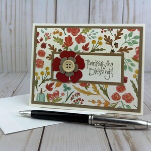 Pu&ograve; includere: Un biglietto d'auguri rettangolare con un motivo di foglie autunnali, fiori e ghiande. Un pannello rettangolare centrale mostra il testo "Thanksgiving Blessings" accanto a un fiore di carta rosso e beige con un bottone centrale color crema.