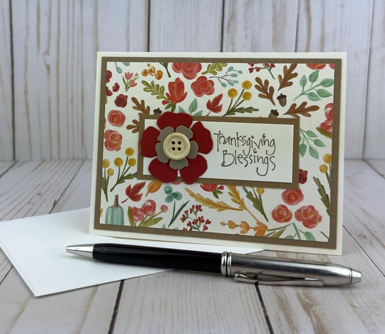 Pu&ograve; includere: Un biglietto d'auguri rettangolare con un motivo di foglie autunnali, fiori e ghiande. Un pannello rettangolare centrale mostra il testo "Thanksgiving Blessings" accanto a un fiore di carta rosso e beige con un bottone centrale color crema.