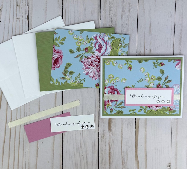 K&ouml;nnte beinhalten: Ein Gru&szlig;kartenset mit hellblauem Hintergrund und rosa-wei&szlig;en Blumenmustern. Die Karte zeigt ein wei&szlig;es rechteckiges Etikett mit dem Text "thinking of you" und drei kleinen klaren Strasssteinen, akzentuiert durch ein schmales cremefarbenes Band.
