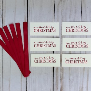 May include: Six rectangular off-white paper tags, each printed with the red text merry CHRISTMAS in a combination of script and block lettering, accompanied by six thin red fabric ribbons arranged in a fan shape to the left of the tags.