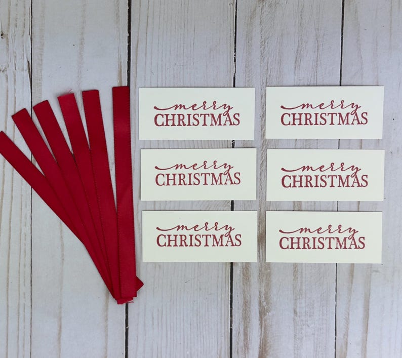 May include: Six rectangular off-white paper tags, each printed with the red text merry CHRISTMAS in a combination of script and block lettering, accompanied by six thin red fabric ribbons arranged in a fan shape to the left of the tags.