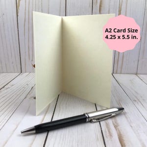 Puede incluir: Una tarjeta A2 plegada de color crema que mide 10,8 x 14 cm, colocada en posici&oacute;n vertical junto a un bol&iacute;grafo negro y plateado. La tarjeta est&aacute; en blanco y presenta una textura lisa, mientras que el bol&iacute;grafo tiene un clip met&aacute;lico y acabado pulido.