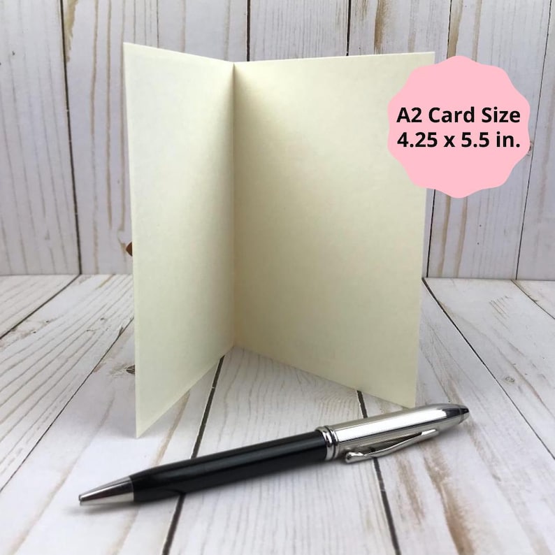 Puede incluir: Una tarjeta A2 plegada de color crema que mide 10,8 x 14 cm, colocada en posici&oacute;n vertical junto a un bol&iacute;grafo negro y plateado. La tarjeta est&aacute; en blanco y presenta una textura lisa, mientras que el bol&iacute;grafo tiene un clip met&aacute;lico y acabado pulido.