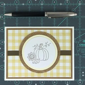 May include: A rectangular greeting card featuring a yellow and white gingham pattern background, a brown horizontal ribbon, and a central circular cutout displaying a black ink line drawing of a pumpkin, a flower, and a leaf. A black pen is placed above.