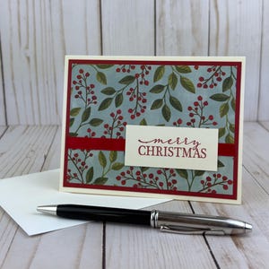 May include: A rectangular greeting card featuring a light blue background with a pattern of green leaves and red berries, framed by a dark red border. A white rectangular label with the red text merry CHRISTMAS is centered on the card.