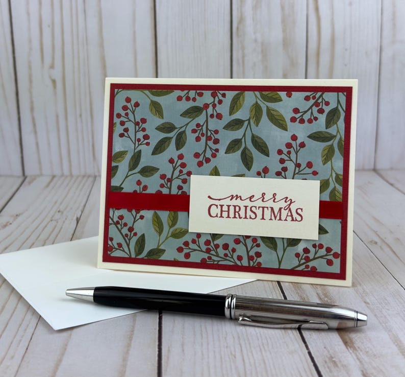 May include: A rectangular greeting card featuring a light blue background with a pattern of green leaves and red berries, framed by a dark red border. A white rectangular label with the red text merry CHRISTMAS is centered on the card.
