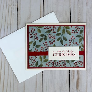 May include: A rectangular greeting card with a light blue background featuring a pattern of green leaves and red berries. A horizontal dark red ribbon crosses the center, topped with a white label displaying the text merry CHRISTMAS.