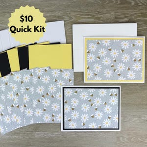 May include: A stationery set featuring cards and envelopes with a light blue background, white daisy pattern, and small yellow and black bees. The kit includes solid yellow and black cardstock inserts and a yellow starburst label reading $10 Quick Kit.