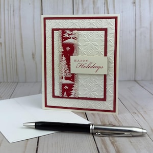 Puede incluir: Una tarjeta de felicitaci&oacute;n de color crema con un patr&oacute;n de copos de nieve en relieve sobre un borde rojo oscuro. Una tira vertical de papel rojo con &aacute;rboles y copos blancos se sit&uacute;a en el centro con la etiqueta Happy Holidays.