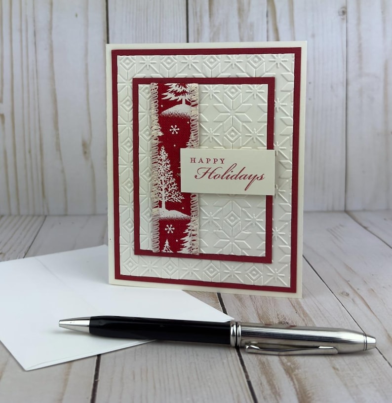 Puede incluir: Una tarjeta de felicitaci&oacute;n de color crema con un patr&oacute;n de copos de nieve en relieve sobre un borde rojo oscuro. Una tira vertical de papel rojo con &aacute;rboles y copos blancos se sit&uacute;a en el centro con la etiqueta Happy Holidays.